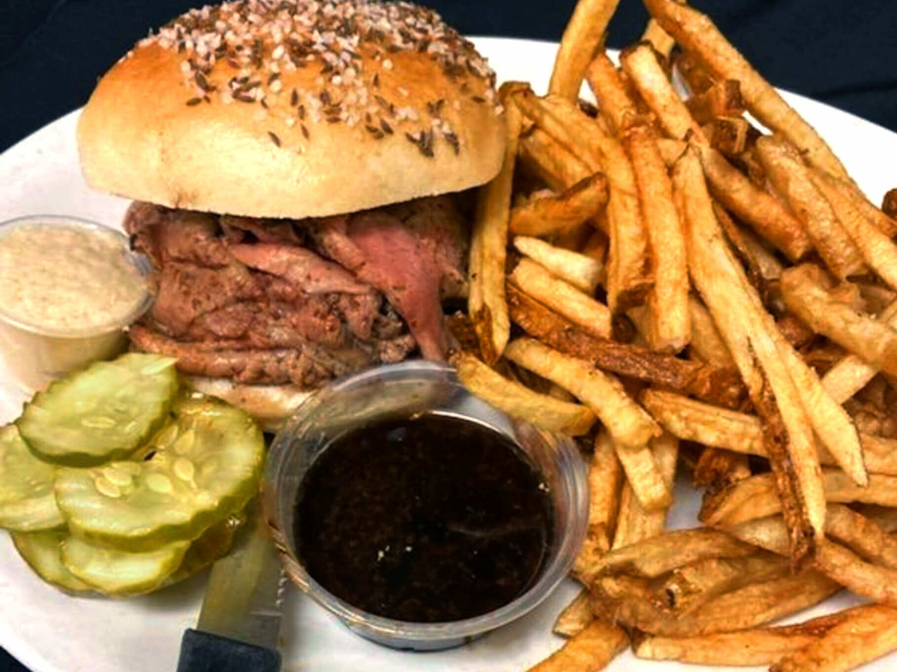 Roast beef sandwich on plate with fries and pickles. 